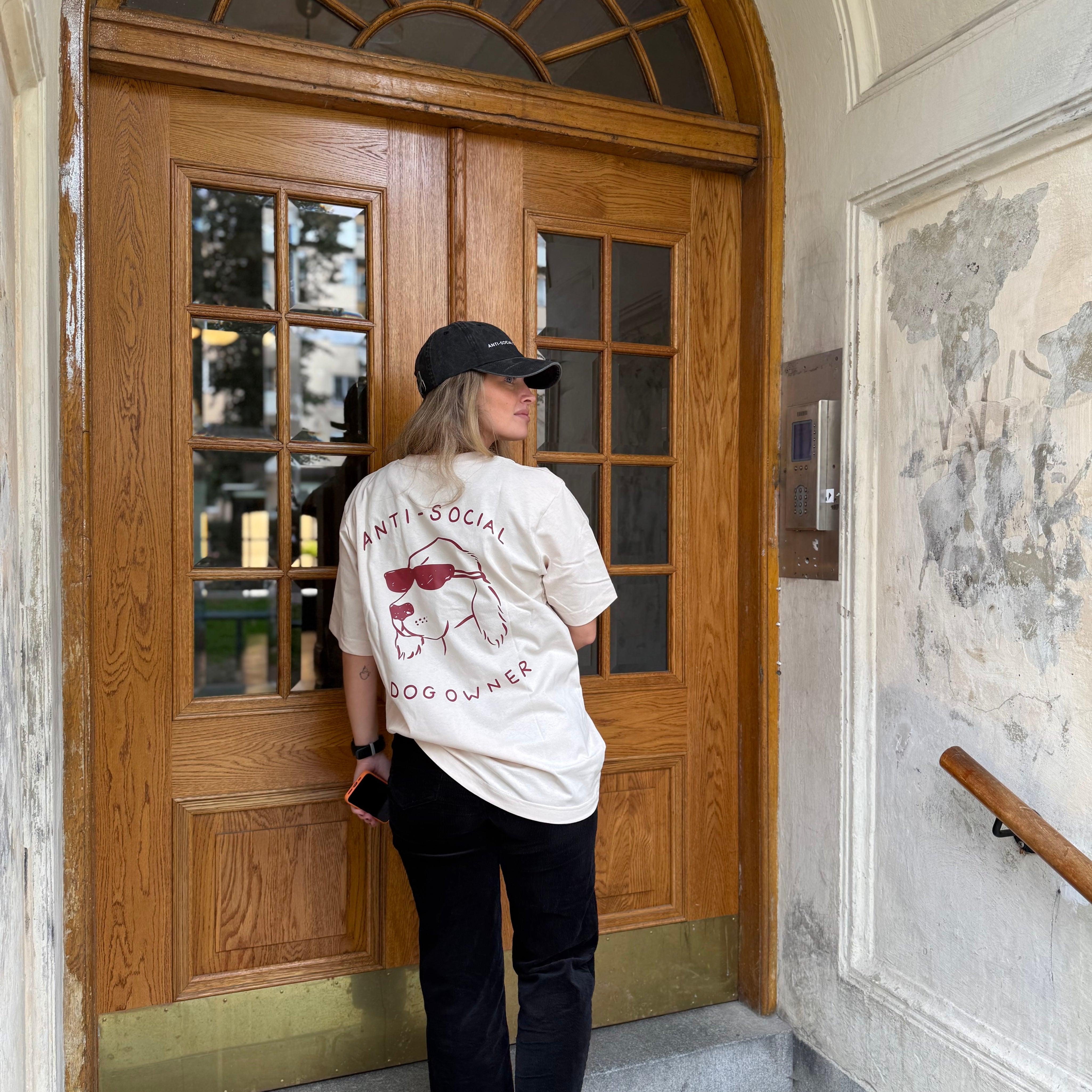 Person wearing a white t-shirt with red text and graphics, black pants, and a black cap standing in front of a wooden door.