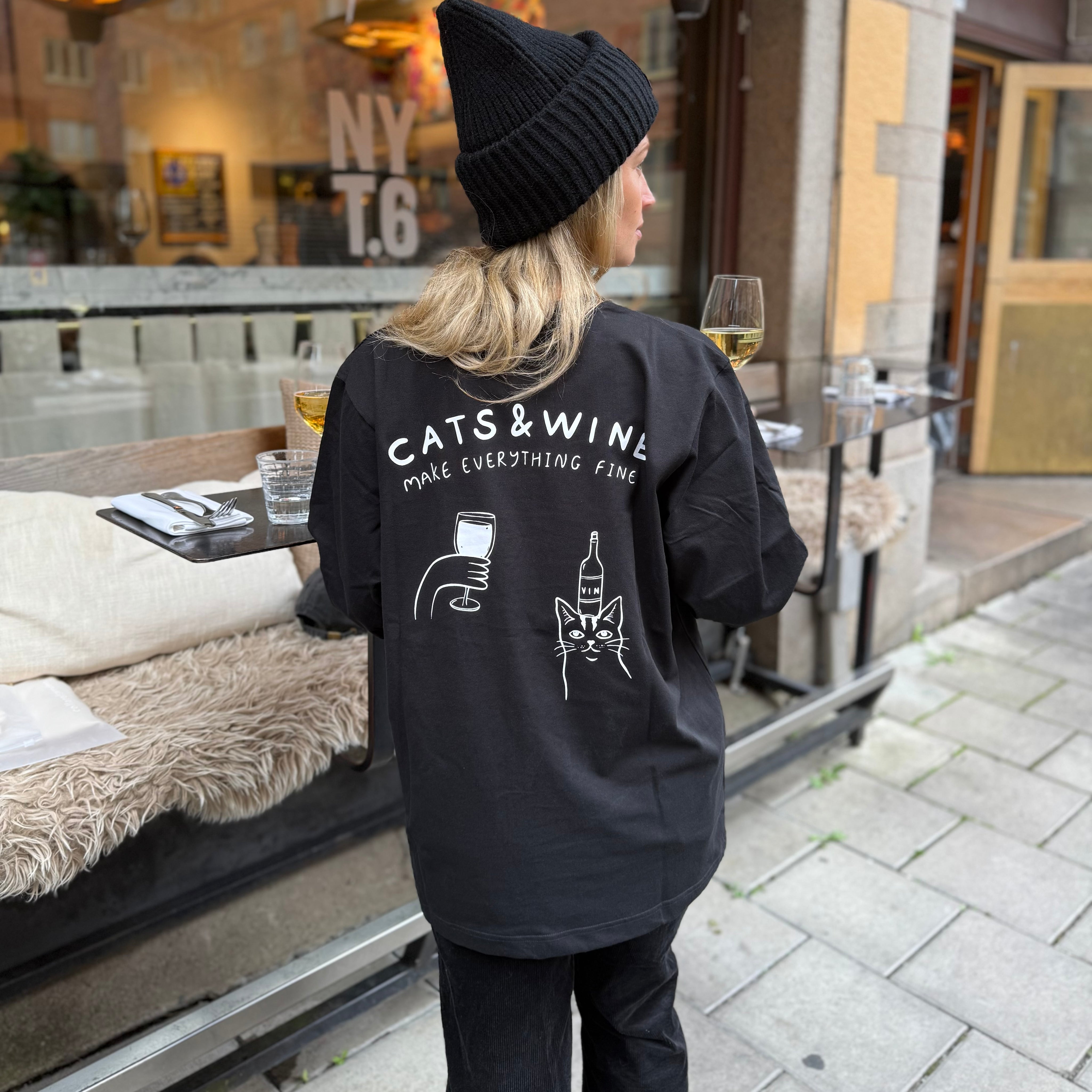 Person wearing a black hoodie with 'Cats & Wine Make Everything Fine' text and graphics, standing outdoors.