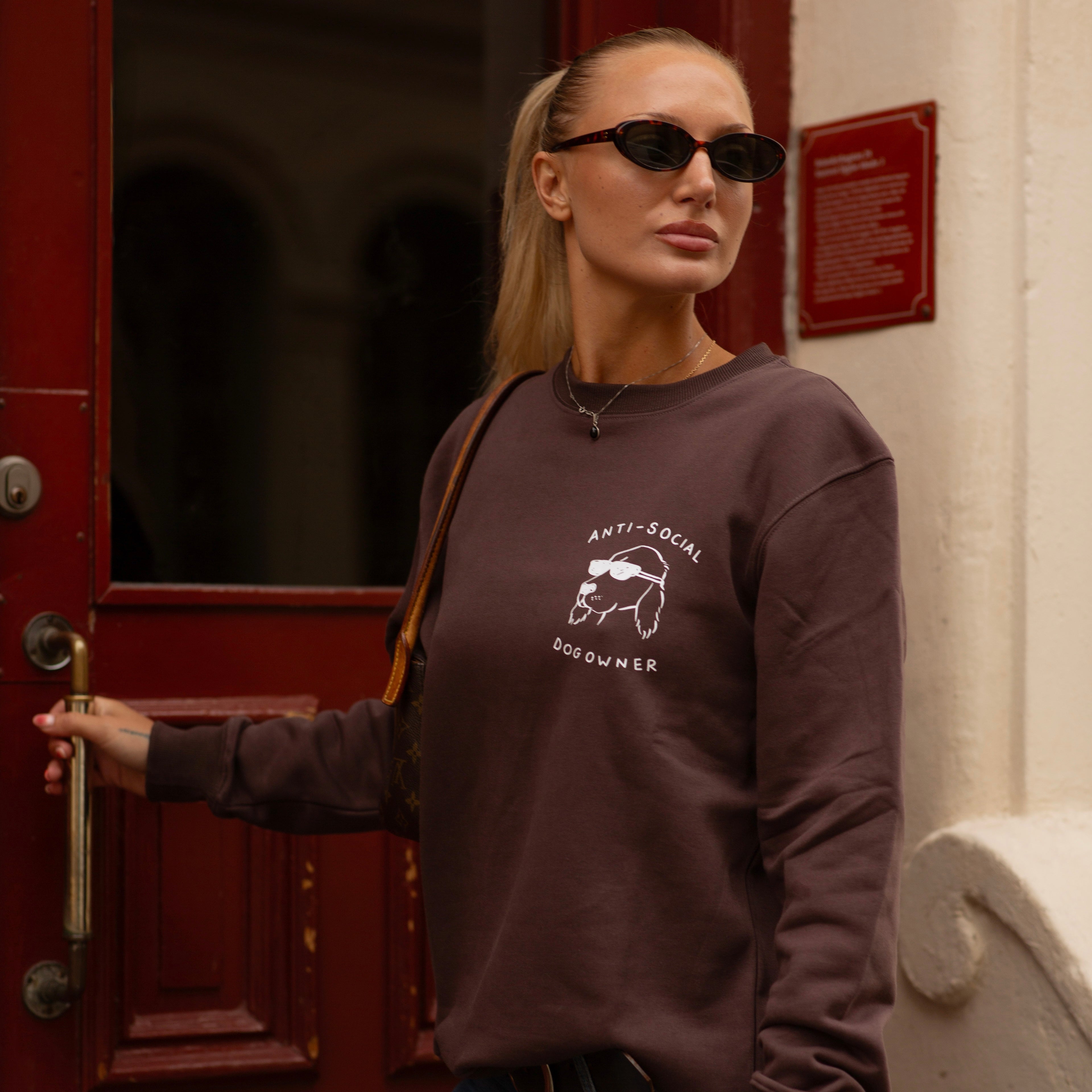 Woman wearing a brown sweatshirt with a graphic design, standing in front of a red door.