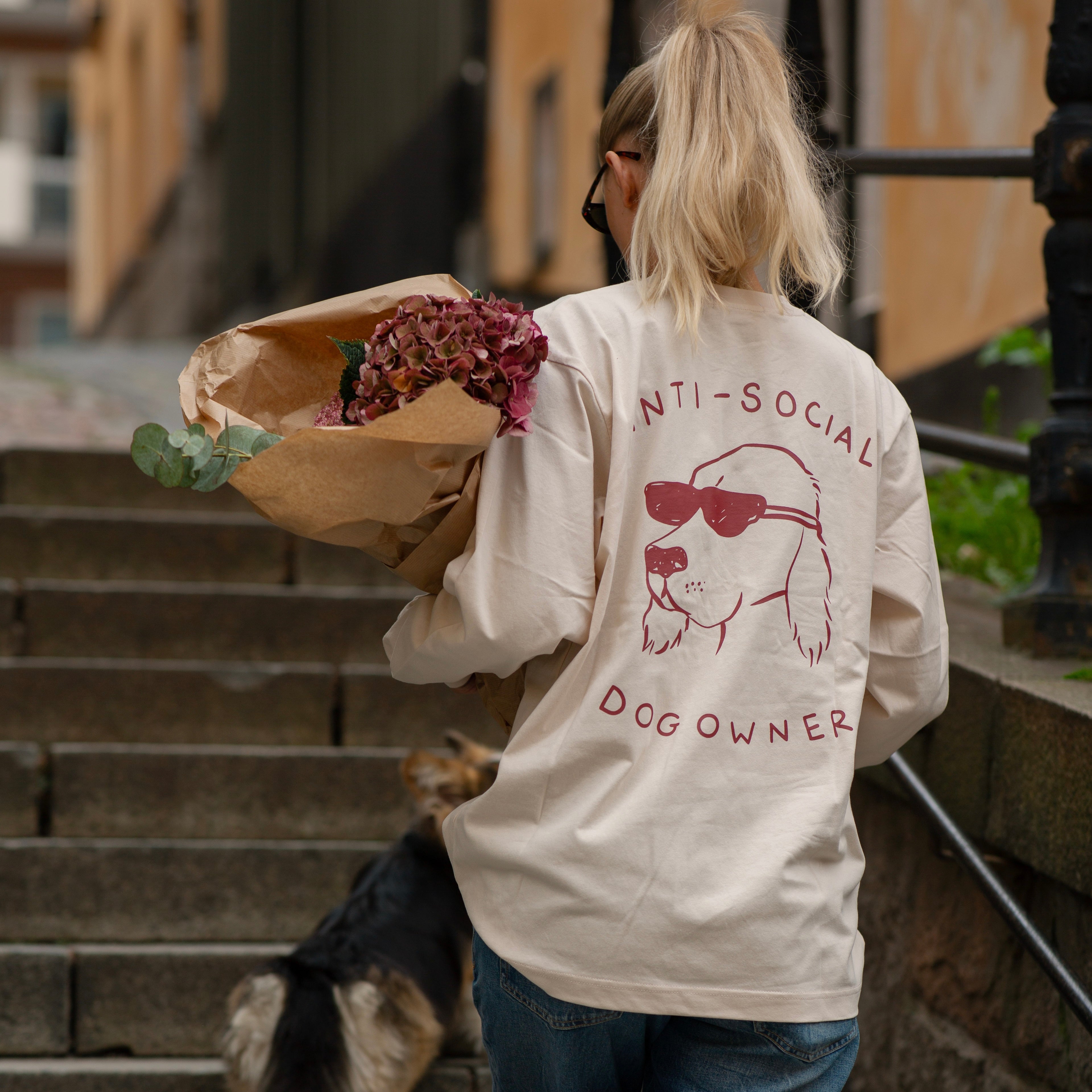 Person wearing a 'ANTI-SOCIAL DOG OWNER' shirt with a dog graphic, holding flowers, walking a dog on a city street.